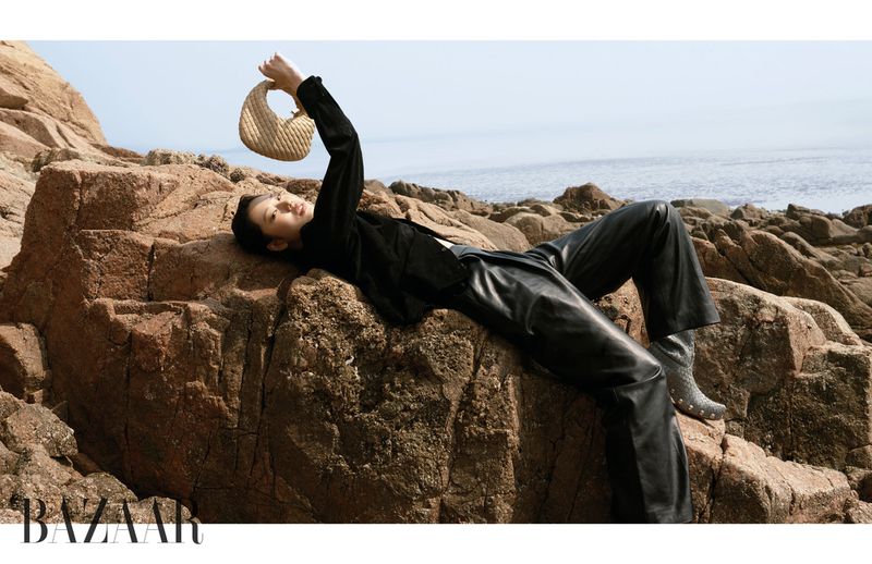 Model Heejung Park reclines on coastal rocks holding a woven beige handbag wearing black leather in Harper's Bazaar Korea.