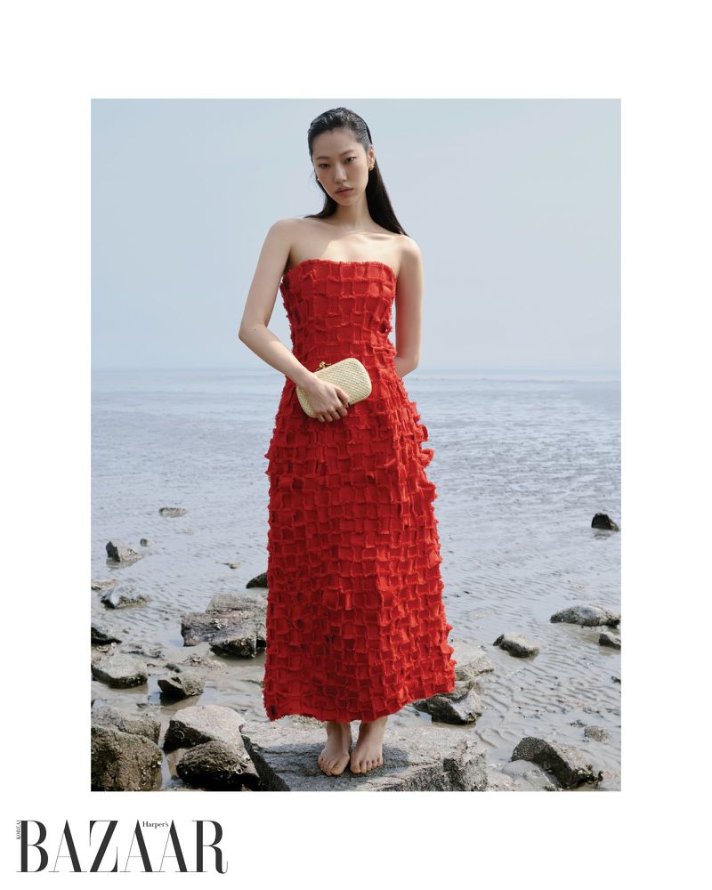Model Heejung Park in a textured red strapless dress holding a clutch while standing barefoot on a rocky shore.