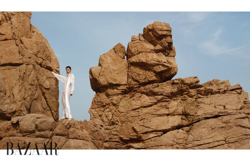 Model Heejung Park in a shimmering white jumpsuit leaning against massive jagged rock formations under a clear sky.