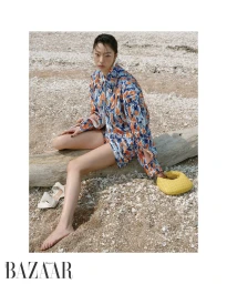 Model seated on a large piece of driftwood on a sandy beach wearing a patterned blue and orange shirt with a yellow handbag.