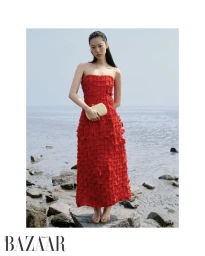 Model Heejung Park in a textured red strapless dress holding a clutch while standing barefoot on a rocky shore.