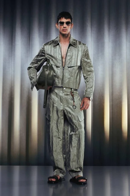 A man in a shiny green outfit poses with a bag in front of a neutral backdrop, wearing sunglasses and sandals in a studio setting.