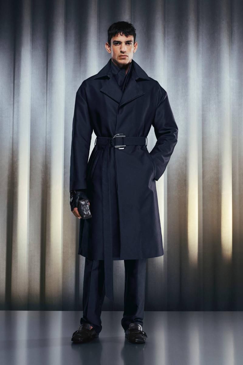 A stylish male model in a long dark trench coat with a belt, standing against a background of vertical silver-toned stripes.