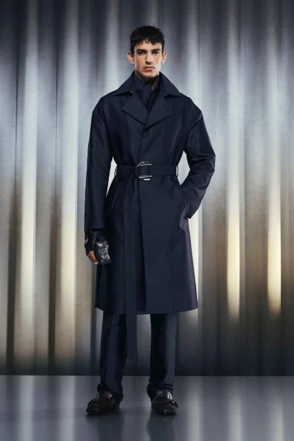 A stylish male model in a long dark trench coat with a belt, standing against a background of vertical silver-toned stripes.