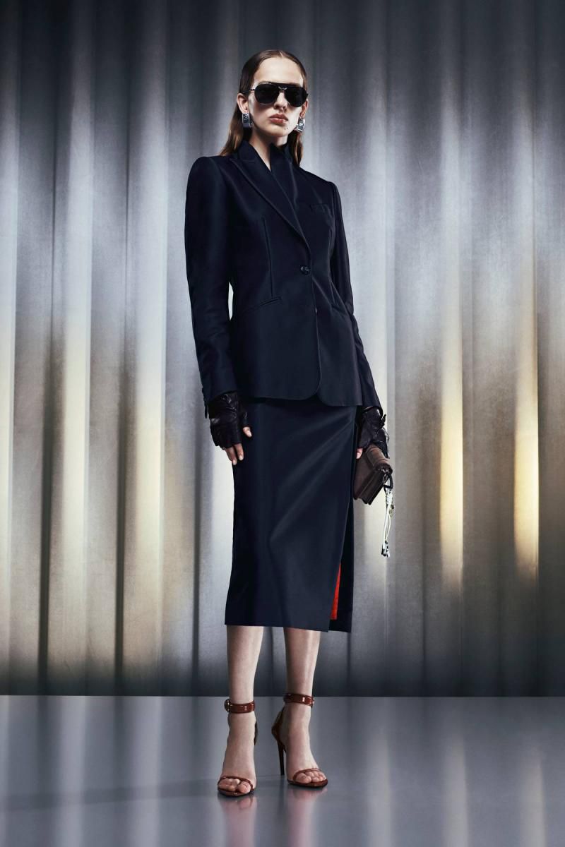 Fashionable woman in black blazer and skirt poses with a handbag against a neutral backdrop, accessorized with sunglasses and heels.