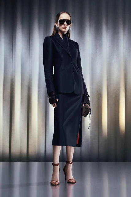 Fashionable woman in black blazer and skirt poses with a handbag against a neutral backdrop, accessorized with sunglasses and heels.