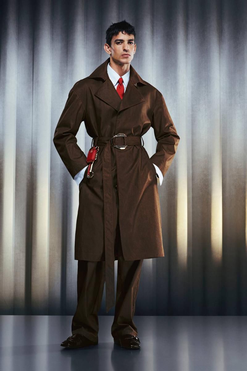 A man in a brown overcoat, white shirt, and red tie poses against a striped backdrop in a full-length shot, showcasing fashion and style.