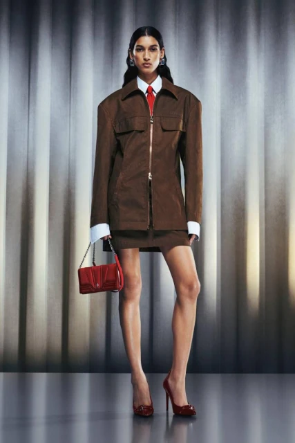 A woman in a brown jacket and skirt stands in a studio holding a red purse, wearing matching red high heels and a red tie.
