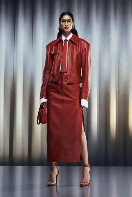 A woman poses in a red leather jacket, matching skirt, and heels against a textured grey backdrop for a fashion editorial shot.