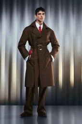 Model wearing a brown belted coat with a white shirt and red tie against a grey textured vertical backdrop
