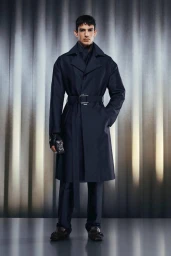 Model wearing a structured dark navy belted trench coat and tailored trousers against a fluted studio backdrop.