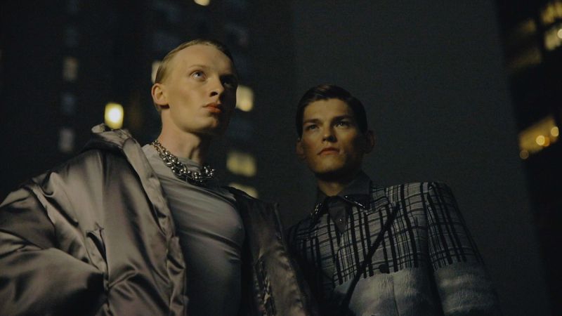 Two adult males model coats with buildings in the background at night, under dramatic lighting, and with a low-angle perspective.