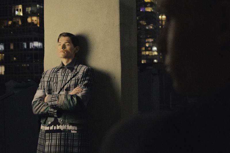 Model leaning against a pillar wearing a grey plaid coat overlooking a city skyline at night with another model in silhouette.