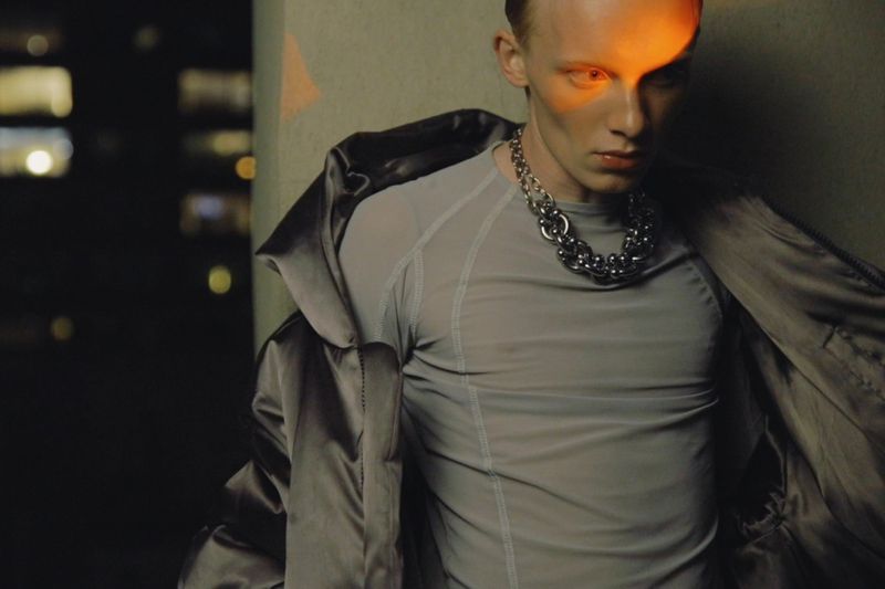 Male model in a grey fitted top and open satin jacket wearing a chunky chain necklace with orange light across his face.