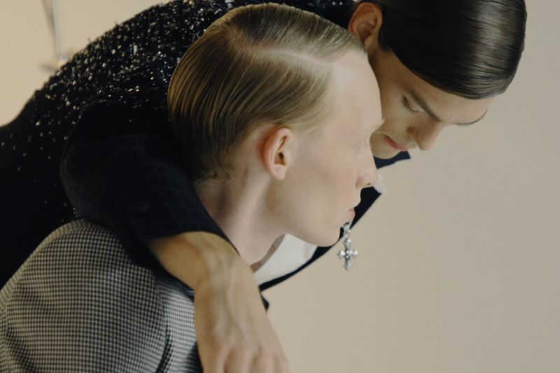 Two male models with slicked-back hair in close embrace, wearing a houndstooth jacket and an embellished black jacket.
