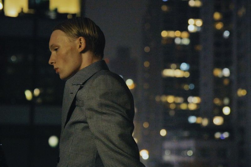 Model in a grey houndstooth blazer standing against a blurred nighttime city skyline with glowing bokeh lights.