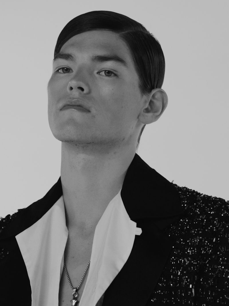 Monochrome portrait of a man with slicked hair wearing an open white collared shirt and embellished black jacket.