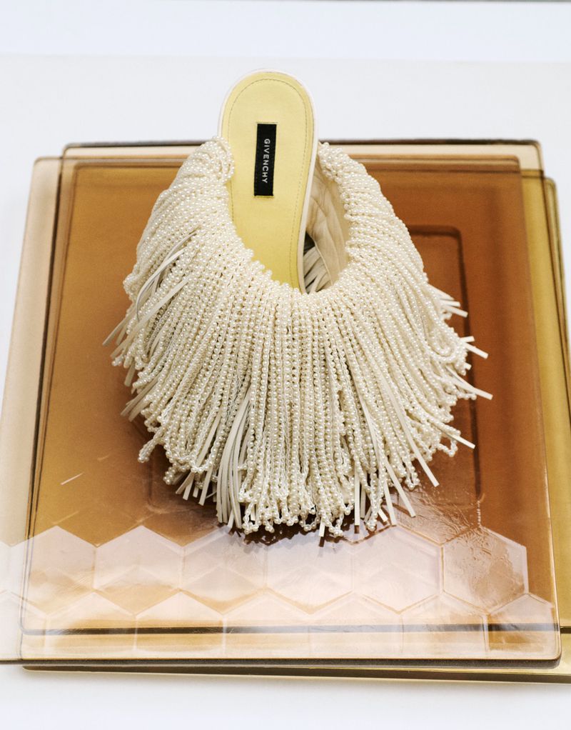 Givenchy off-white beaded fringe slip-on heel shoe displayed on amber glass trays against a bright background.