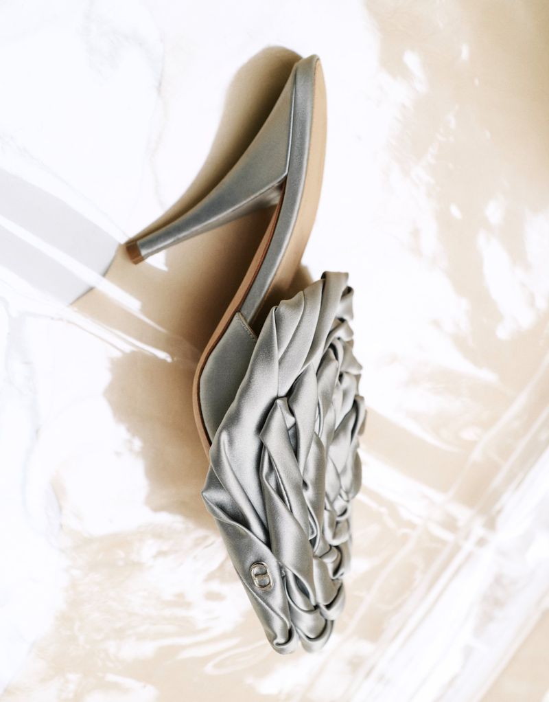 Silver satin pointed-toe stiletto mule with ruched folded fabric detailing resting on a glossy reflective plastic sheet.