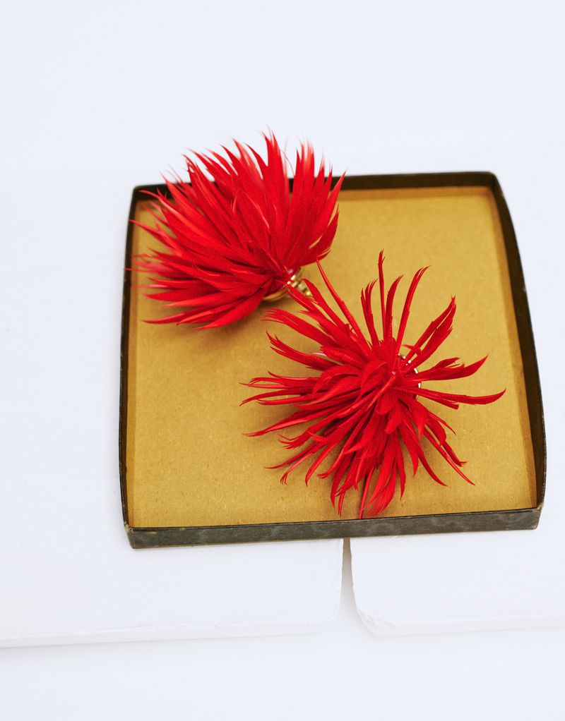 A pair of spiky red feather flower earrings presented inside an open tan and black jewelry box on a white surface.