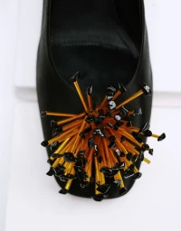 Top-down close-up of a black satin shoe adorned with a sculptural burst of gold rods and black beads against a plain white background.