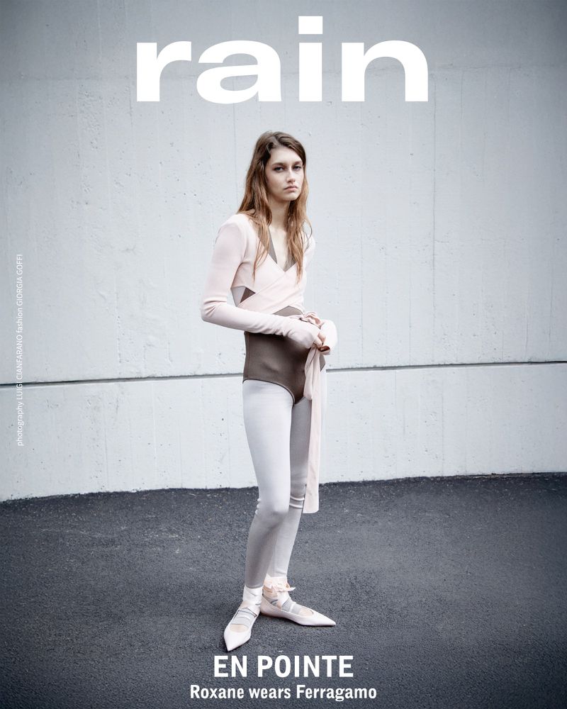 A fashion editorial shot with a woman wearing a leotard, tights, and ballet slippers, posing against a neutral urban backdrop. The magazine name 'rain' is visible.