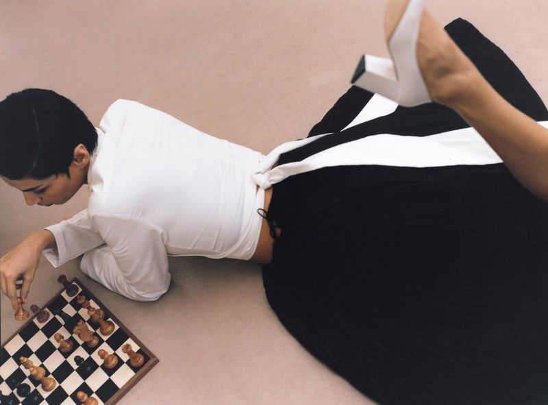 A woman with short hair lying on the floor in a white wrap top and voluminous black skirt playing a game of chess.