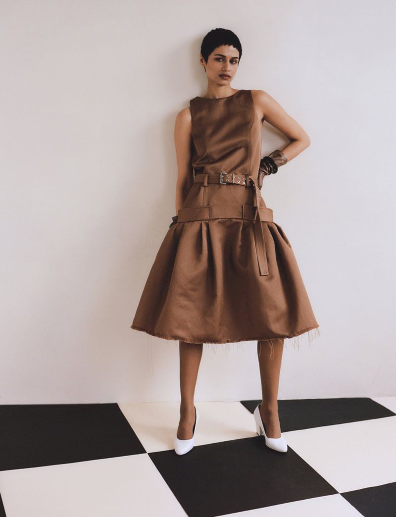 Model in brown belted sleeveless dress and white high heels standing on a black and white checkerboard floor