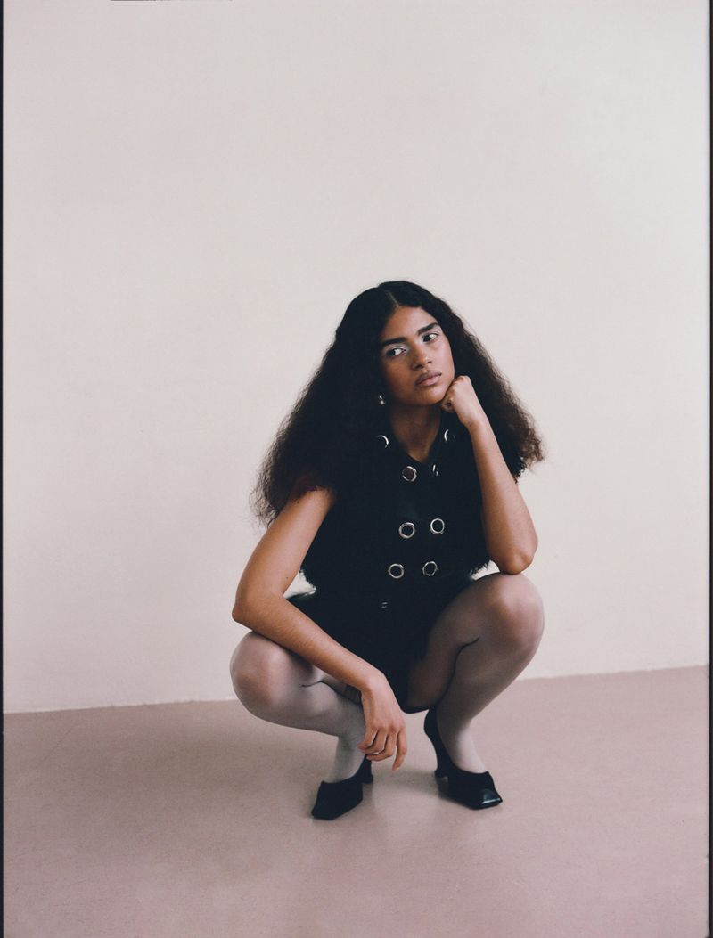 Model Kirandeep Chahal crouching in a black eyelet top, sheer tights, and square-toe heels in a studio setting.