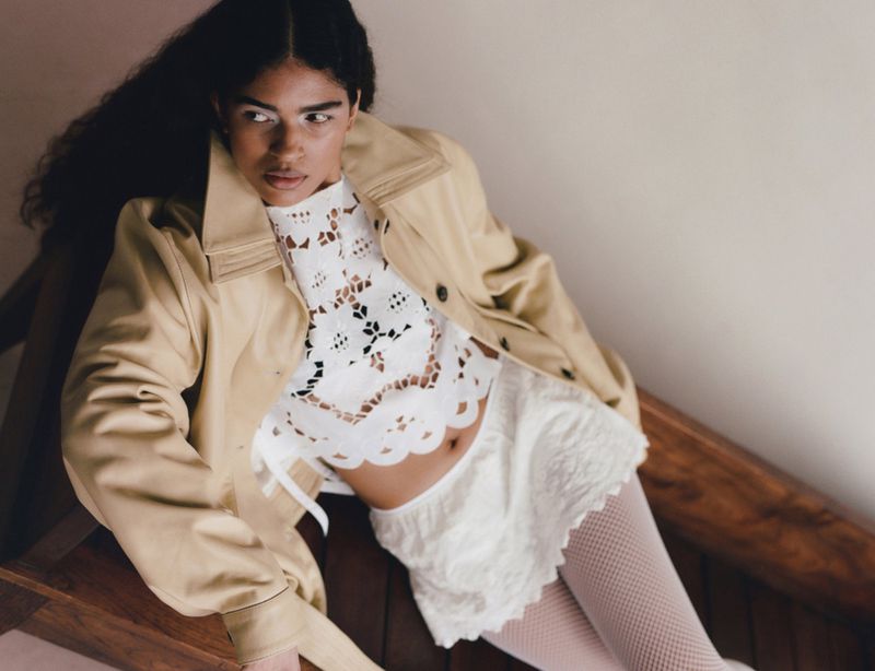 Kirandeep Chahal reclining in a tan trench coat, white cutout lace crop top, and lace mini skirt on a wooden chair.
