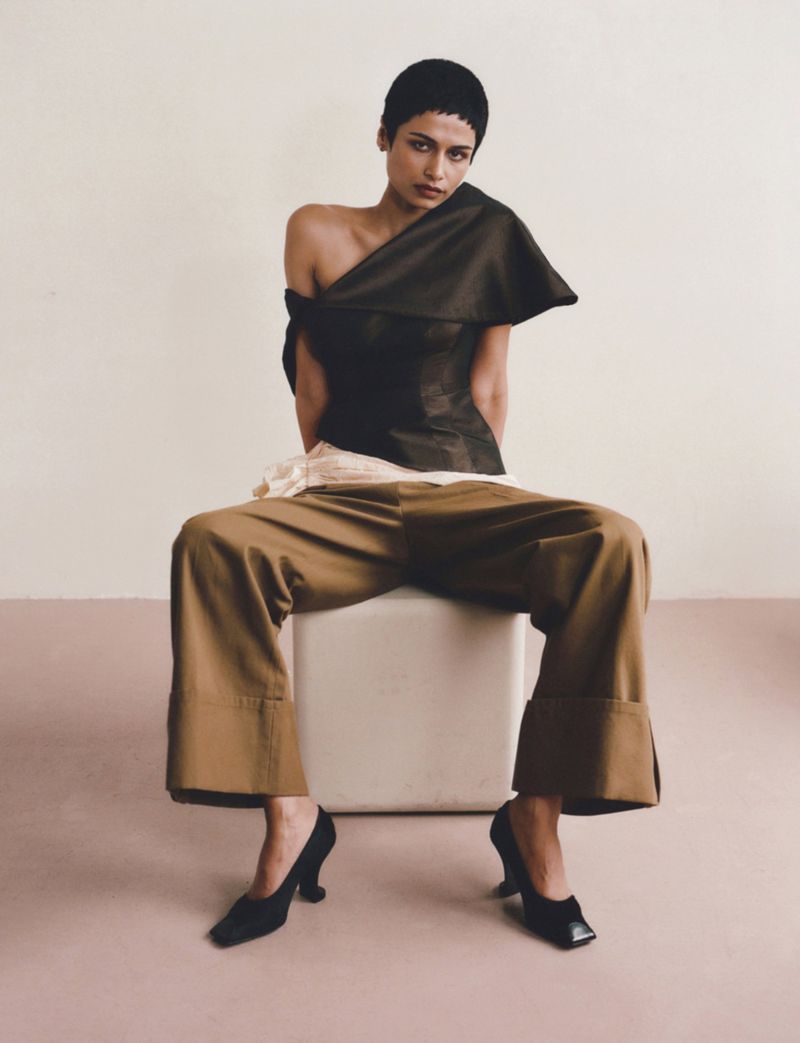 Model with short dark hair seated on a white cube wearing an asymmetrical dark top, wide-leg camel trousers, and black heels.