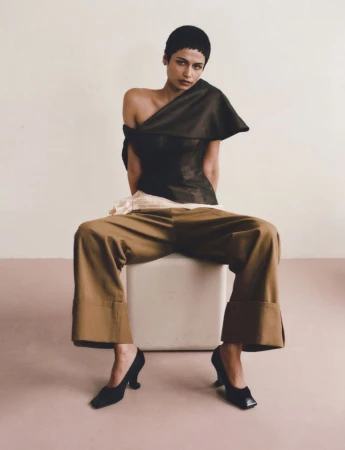 Model with short dark hair seated on a white cube wearing an asymmetrical dark top, wide-leg camel trousers, and black heels.