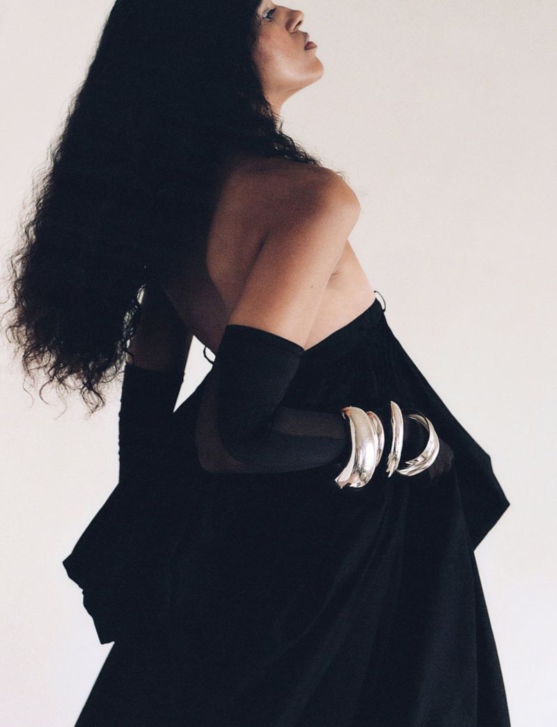Model wearing a black strapless gown, sheer black gloves, and silver sculptural bangles against a white studio background.