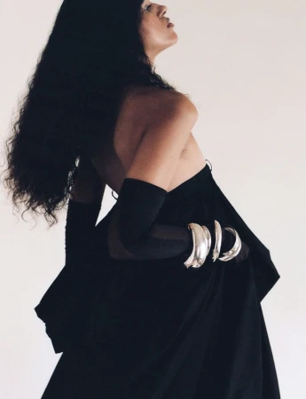 Model wearing a black strapless gown, sheer black gloves, and silver sculptural bangles against a white studio background.