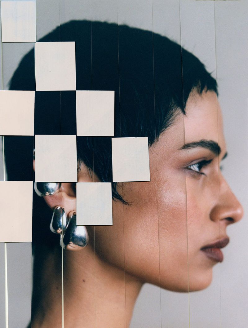 Profile portrait of a woman with short hair wearing silver sculptural earrings, overlaid with white square tiles and vertical line cuts.