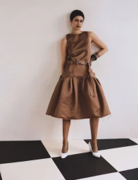 Model in brown belted sleeveless dress and white high heels standing on a black and white checkerboard floor
