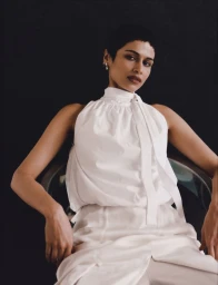Model Kirandeep Chahal poses seated wearing a gathered white sleeveless high-neck top against a black seamless backdrop.