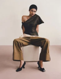 Model with short dark hair seated on a white cube wearing an asymmetrical dark top, wide-leg camel trousers, and black heels.