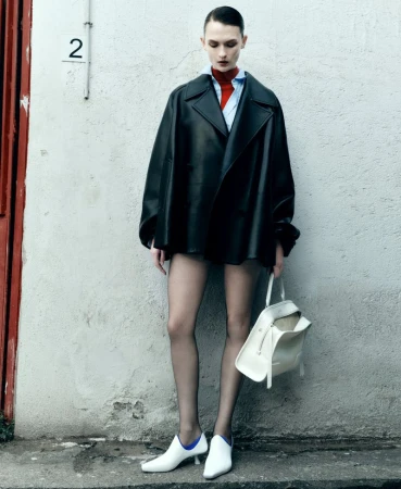 Model Lara Mullen posing in an oversized black leather coat, sheer tights, and white heels against an exterior stucco wall.