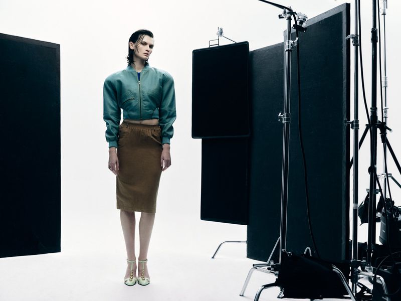 Model Lara Mullen in a cropped teal leather jacket and brown pencil skirt on a studio set with lighting grip equipment.