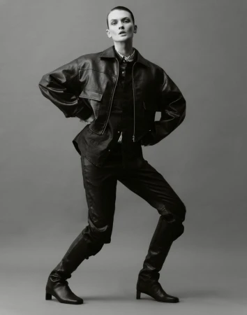 Full-length portrait of a fashion model in a black leather jacket, pants, and boots against a grey background in a studio setting.