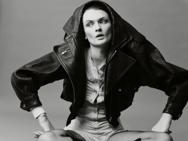 Monochrome portrait of a model in a black hooded leather jacket and light-colored shorts, seated with hands on thighs in photo studio.
