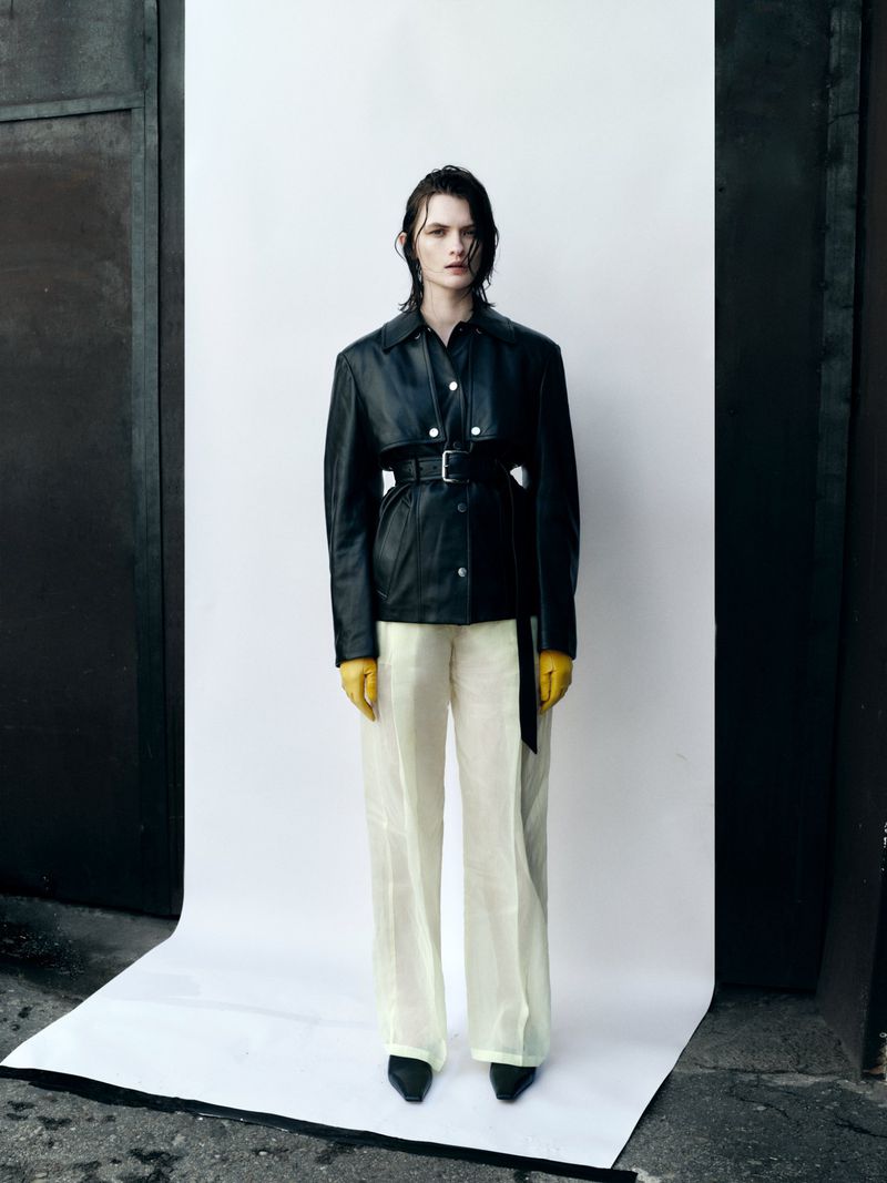 Model Lara Mullen wearing a belted black leather jacket, yellow gloves, and cream trousers against a white seamless backdrop.