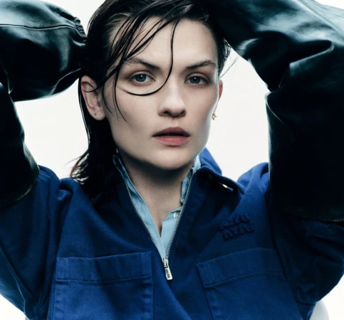 Close-up portrait of a woman with wet-look hair wearing a blue zip-up jacket and black leather sleeves against a stark white background.