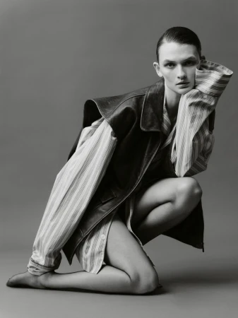 Model Lara Mullen crouching in an oversized striped shirt and dark leather vest against a studio backdrop in black and white.