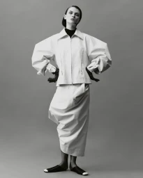 Model Lara Mullen posing in a sculptural white balloon-sleeve jacket and long draped skirt against a grey studio backdrop.