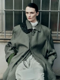 Model Lara Mullen seated in an oversized textured sage coat and white trousers before a barred window.