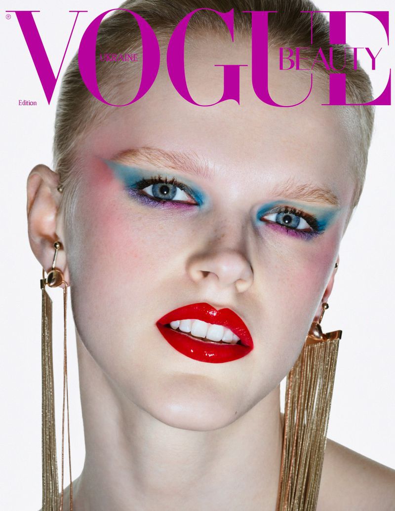 Close-up of a woman with colorful eyeshadow, red lipstick, and long gold earrings on a bright white background, Vogue magazine cover.