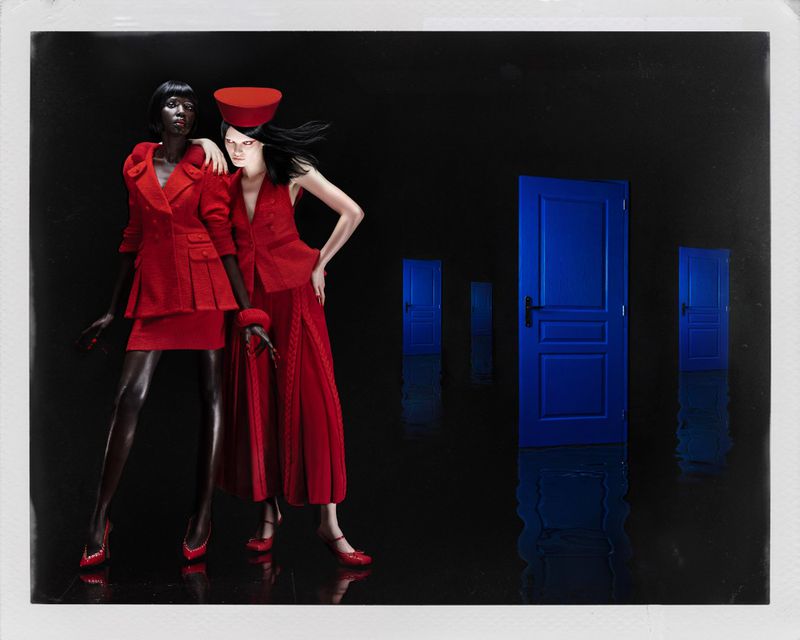 Two fashion models wearing red outfits, posing in front of blue doors on a black background in a studio-style shot.