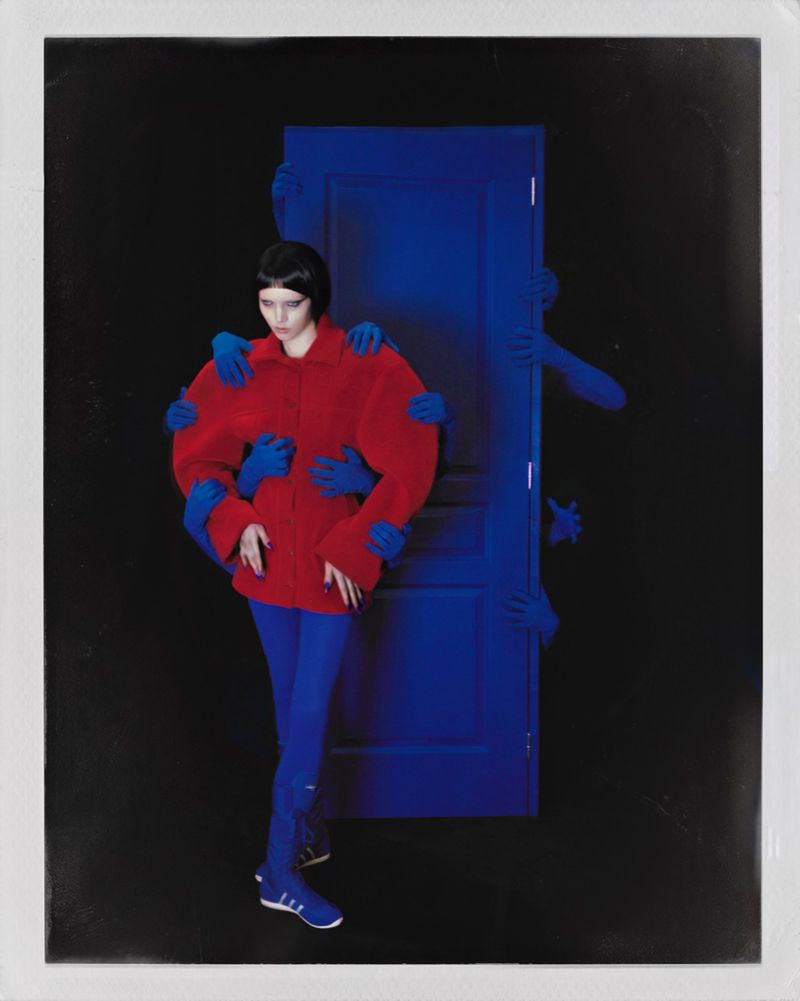 Fashion model in an oversized red sculptural coat and blue tights standing before a blue door with reaching blue hands on a black background.